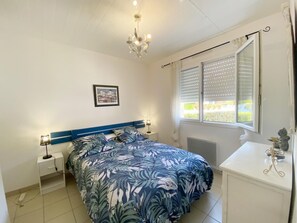 Une chambre à coucher comprenant un lit, des tables de chevet, une commode, une fenêtre avec des stores, un radiateur et un lustre.