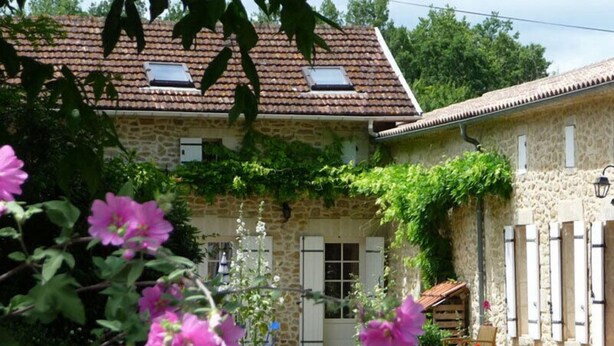 Una casa de pedra amb teulada de teules, un jardí petit amb flors roses i una boscadeira de fusta.