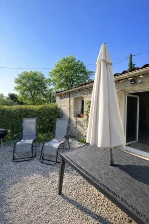 Une terrasse avec deux chaises longues et un parasol blanc, un bâtiment en pierre avec une porte, et une haie en arrière-plan.
