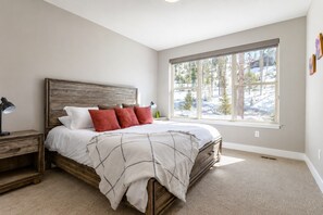 A bedroom with a large bed, a nightstand, a window with a view of trees, and a wall with a picture.