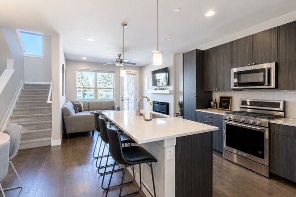 A modern kitchen with a central island, dark cabinetry, stainless steel appliances, and a staircase leading to a living area.