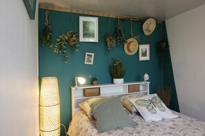 A bedroom with a bed, a bedside lamp, a shelf with plants, and a wall with hanging plants and hats.
