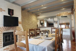 A dining area with a stone wall, a fireplace, wooden chairs, a table set for four, and a kitchen visible in the background.