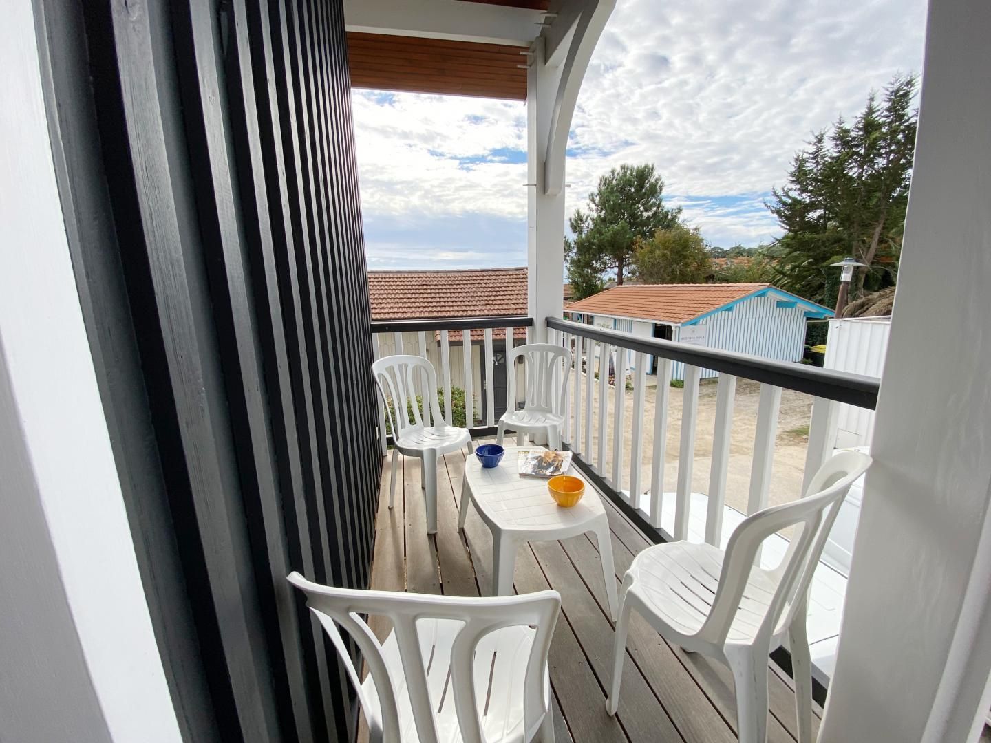 Un balcon avec des chaises en plastique blanches et une petite table, donnant sur un quartier résidentiel avec des maisons et des arbres.