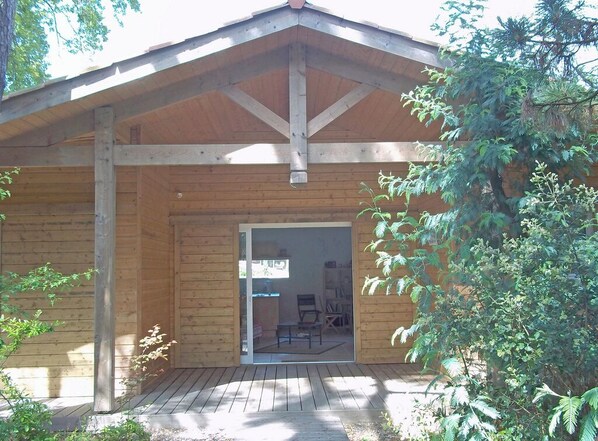 A wooden cabin with a covered porch, a sliding glass door leading to an interior with a desk and chair, surrounded by trees.