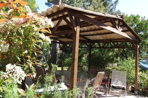 Une pergola en bois, avec une table et des chaises en dessous, entourée de verdure et d’arbres.