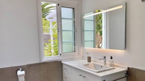 Een badkamer met een wit wastafel, spiegel, en kastje, een toiletrolhouder, en een raam met uitzicht op groen.