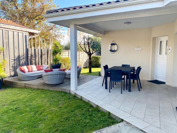 Un patio cubierto con una mesa de comedor y sillas, una zona de estar con cojines y un pequeño jardín.
