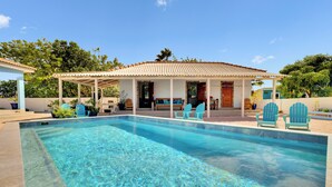 A swimming pool with a covered patio area, outdoor seating, and a view of a house with a tiled roof.