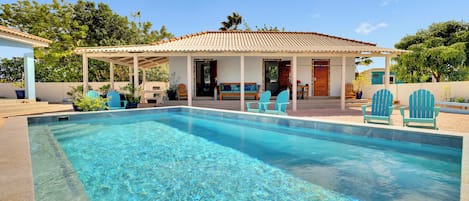 A swimming pool with a covered patio area, outdoor seating, and a view of a house with a tiled roof.