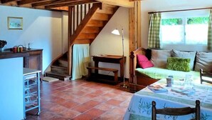 Un interior rústico con vigas de madera a la vista, una escalera, un sofá con cojines, una mesa con mantel y una ventana con cortinas.