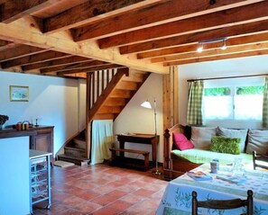 A rustic interior with exposed wooden beams, a staircase, a sofa with cushions, a table with a tablecloth, and a window with curtains.