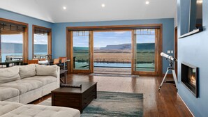 Un salón amplio con un sofá grande, una mesa de centro y una chimenea. Hay ventanas grandes que ofrecen vistas a un lago y montañas.