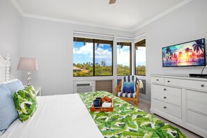 Ein Schlafzimmer mit Bett, Sessel, Fernseher und Fenster mit Blick auf Palmen.