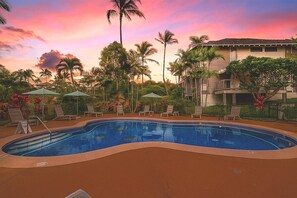 Une piscine entourée de palmiers et d’un bâtiment doté de balcons, avec des chaises longues et des parasols à proximité.
