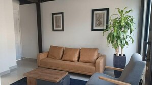 A modern living room with a brown leather sofa, a wooden coffee table, a grey armchair, and a large potted plant. - Study area Artz Pedregal, Hospital Ángeles, IPPC (Ciudad de México)