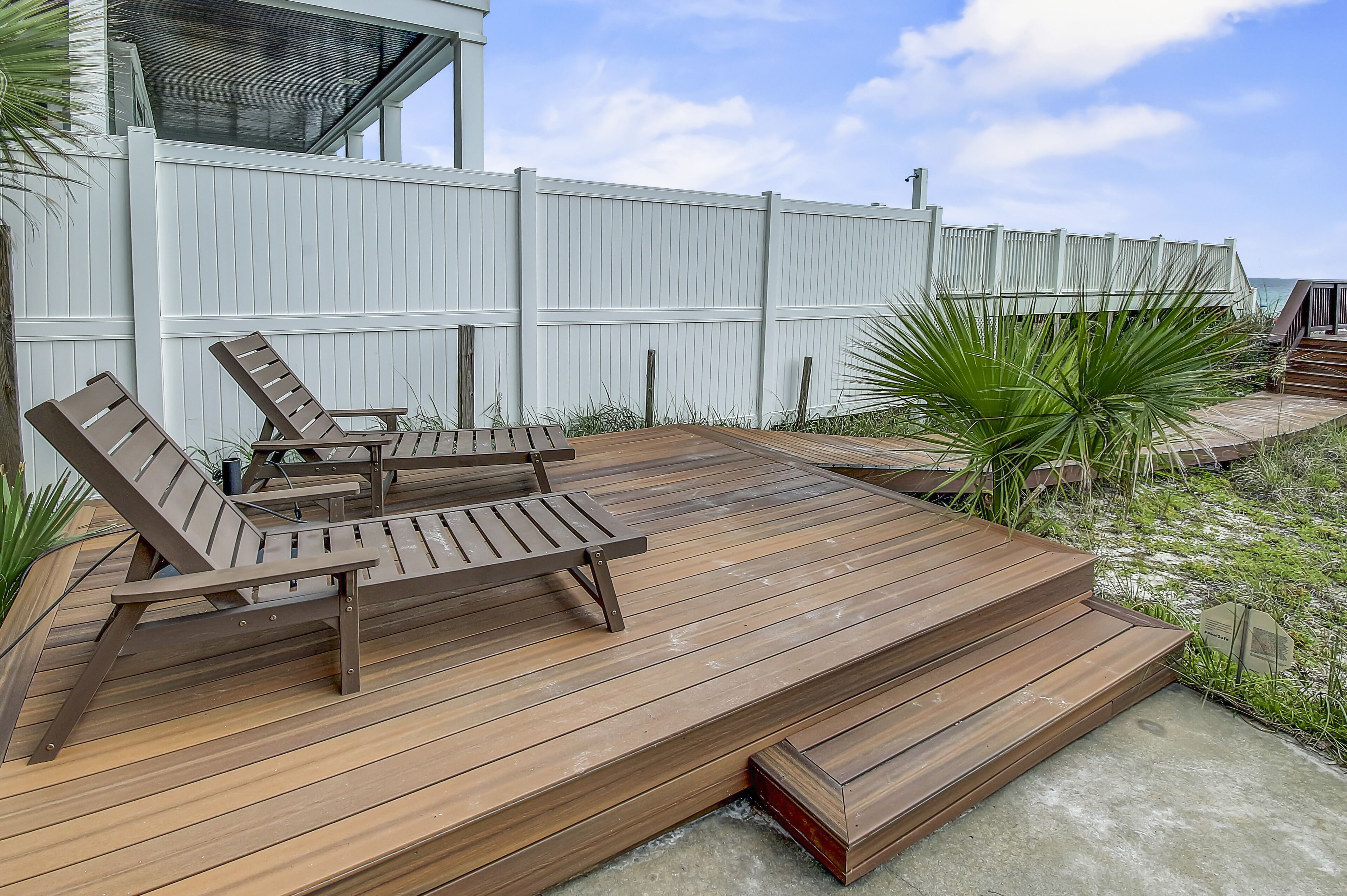 A wooden deck with two lounge chairs and a small table, surrounded by a white fence and tropical plants.