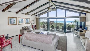 A spacious living room with a large window overlooking the beach, a comfortable sofa, a television, and a bookshelf. - Gulf Front 3bd/2ba Home With Private Beach Access! (Panama City Beach)