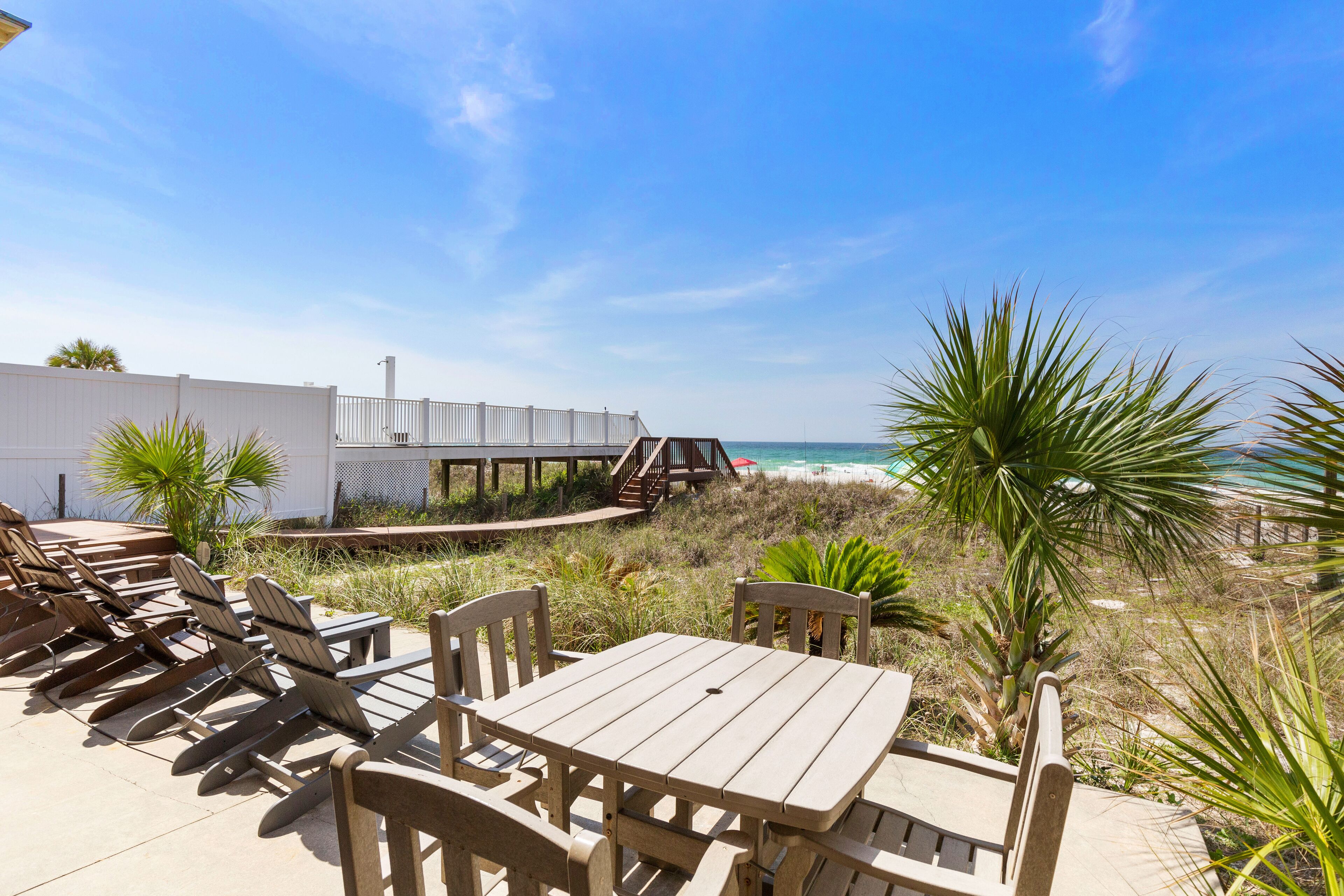 A wooden deck with lounge chairs and a table, overlooking a beach with a white fence and palm trees.