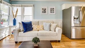 A living room with a beige sofa, a coffee table, and a modern kitchen with a stainless steel refrigerator. - New Jersey Abode - Near Theme Parks, Walk to Beach (Keansburg)
