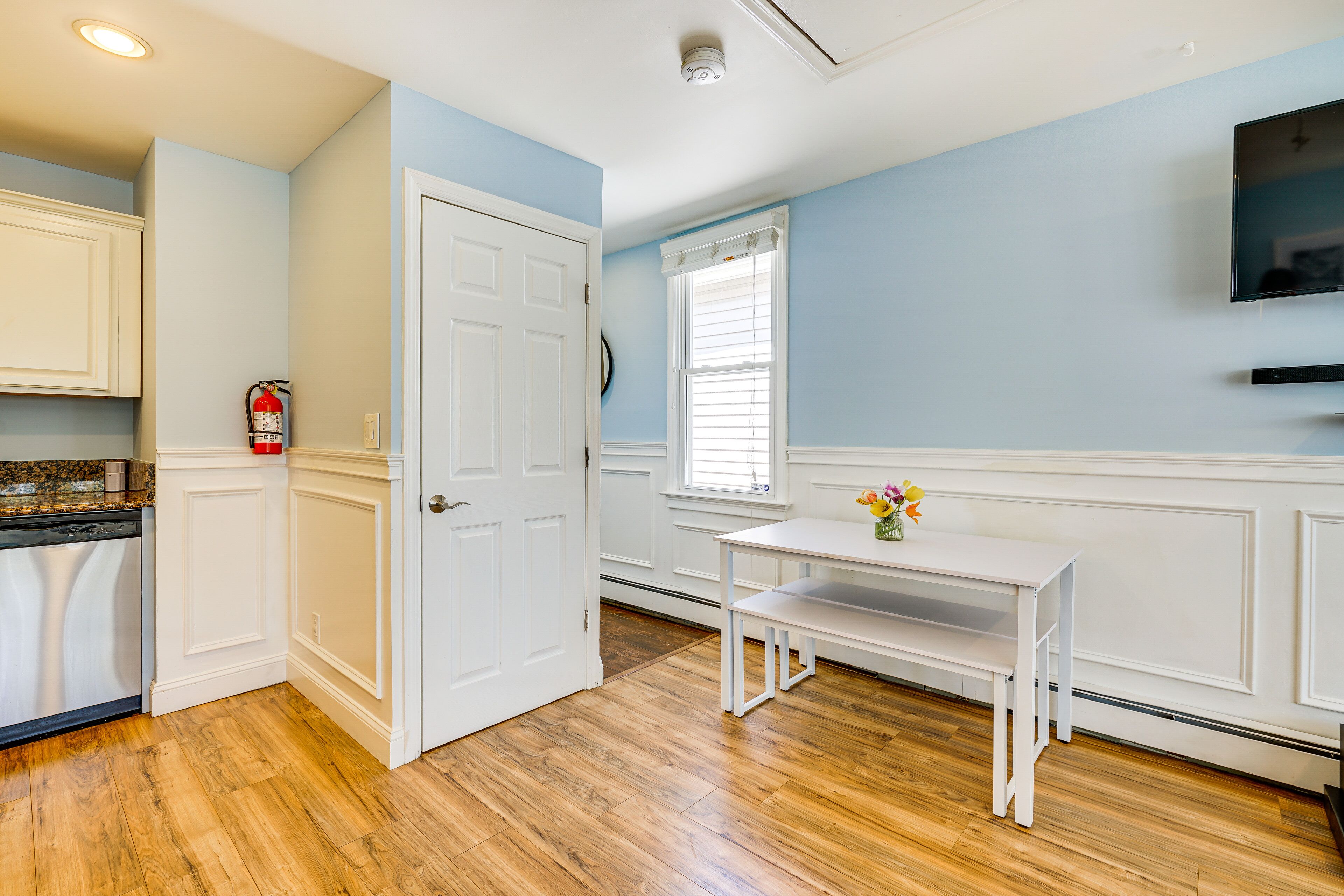 A kitchen with white cabinetry, a wooden floor, a white door, a window with blinds, and a small table with a vase of flowers.