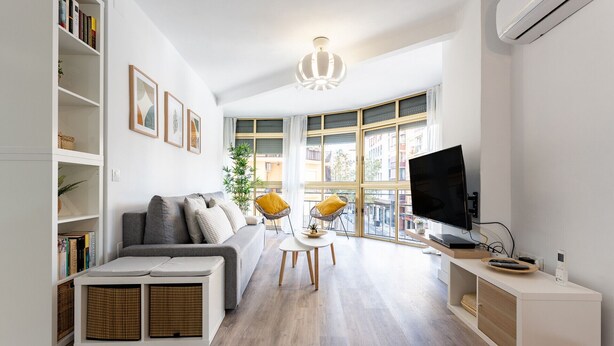 Ett modernt vardagsrum med en grå soffa, ett litet vitt bord och en tv som står på ett tv-bord. På vänster sida finns bokhyllor och ett stort fönster med utsikt över byggnaderna utanför.