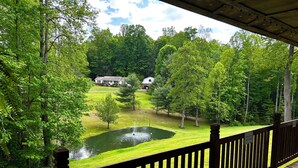 A wooden deck overlooking a pond with a fountain, surrounded by lush greenery and a house in the background.
