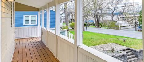 A covered porch with a wooden floor and white railings, overlooking a residential street and a grassy area.