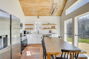 Une cuisine moderne avec des meubles blancs, une table à manger en bois et une vue sur l’extérieur grâce à de grandes fenêtres.