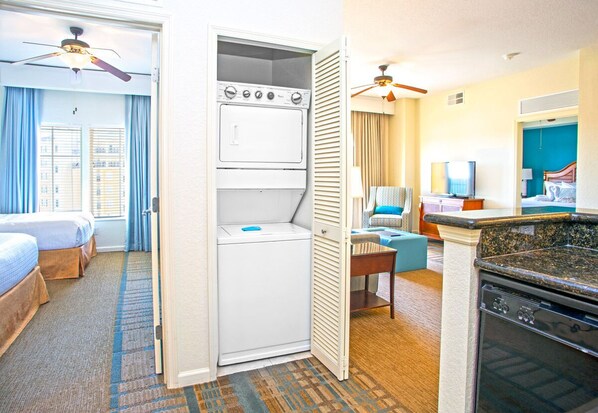 A hotel room with a bed, a washing machine, a dining area with a table and chairs, and a kitchen with a stove.