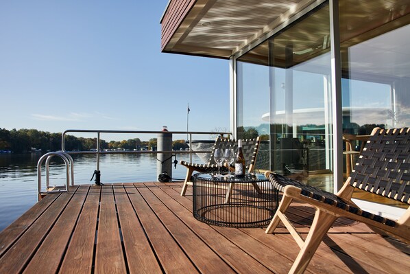 A wooden deck with two lounge chairs and a table, overlooking a calm body of water with a bridge in the background. - Spreeapartment JULIA (Berlin)