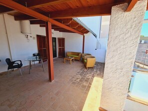 A covered patio area with tiled flooring, a table with chairs, wicker furniture, and a white wall with a window and a door.