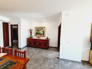 A dining area with a wooden table and chairs, a marble floor, a wooden cabinet with a painting, and a door leading to another room.