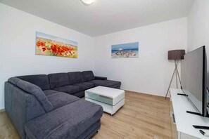 Un salon moderne avec un canapé d’angle gris, une table basse blanche et un téléviseur à écran plat posé sur un meuble blanc.