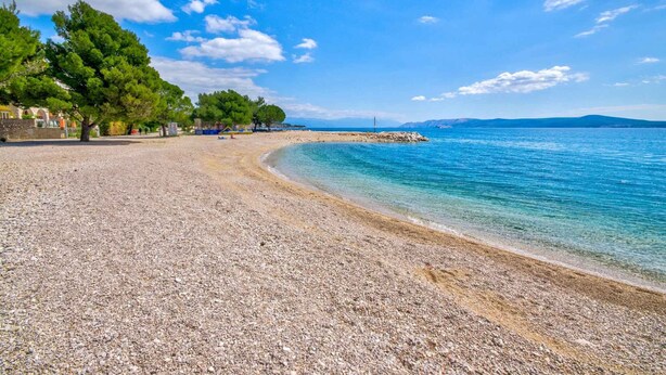 Une plage de galets bordée d’une eau d’un bleu limpide, avec des arbres et une côte visible au loin.