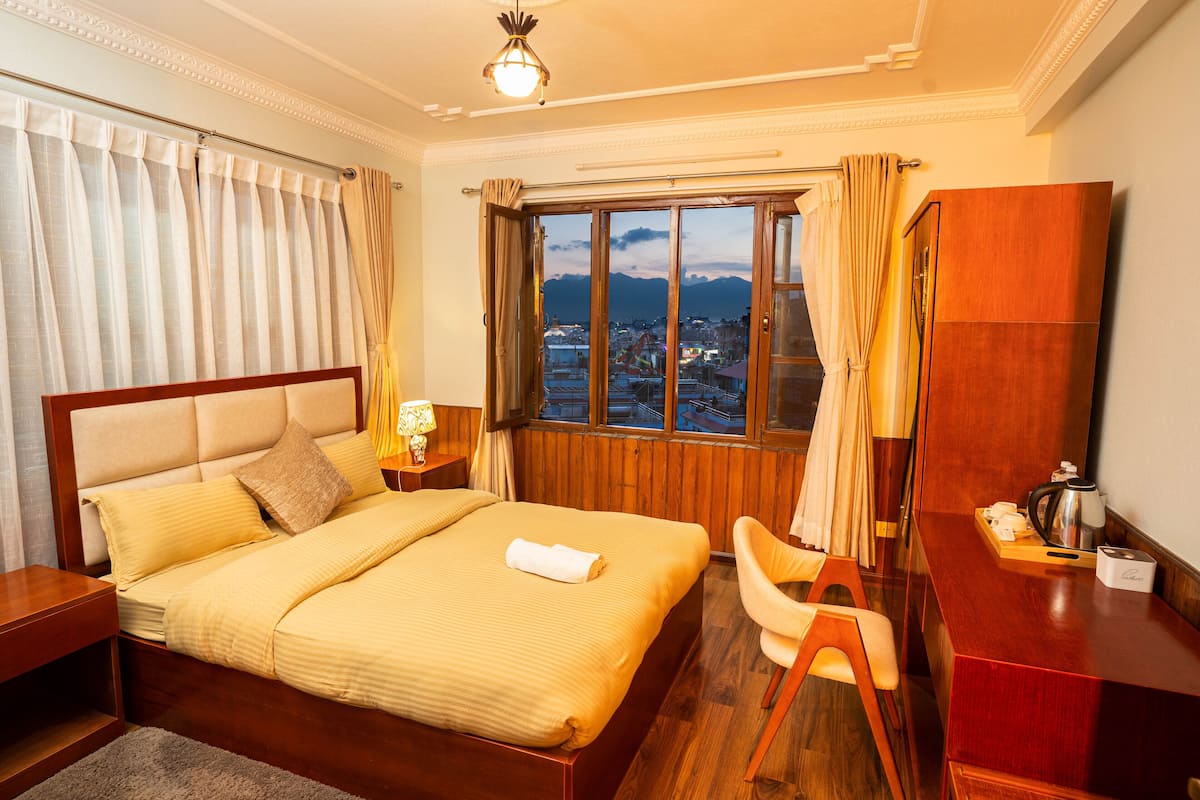 A hotel room with a large bed, a wooden desk, a chair, a mirror, and a window with a city view.