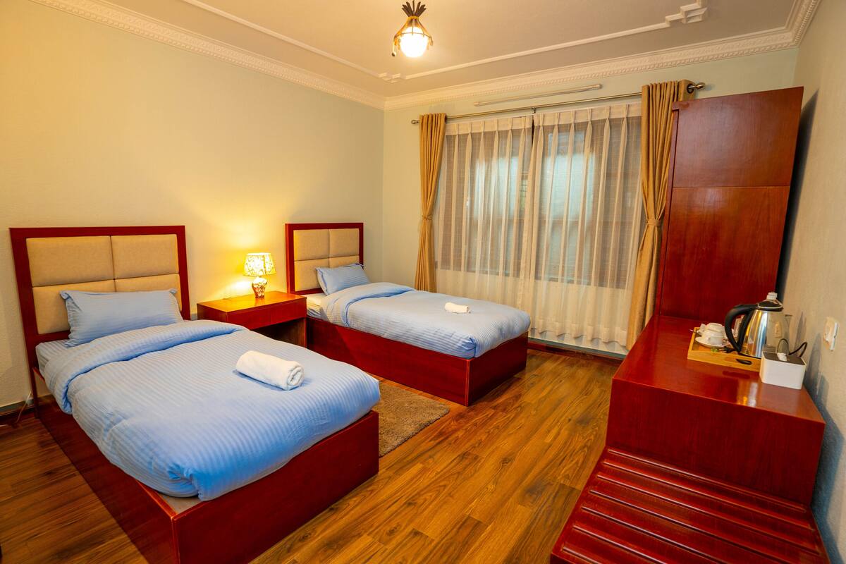 A hotel room with two beds, wooden flooring, a desk, a lamp, and a window with curtains.