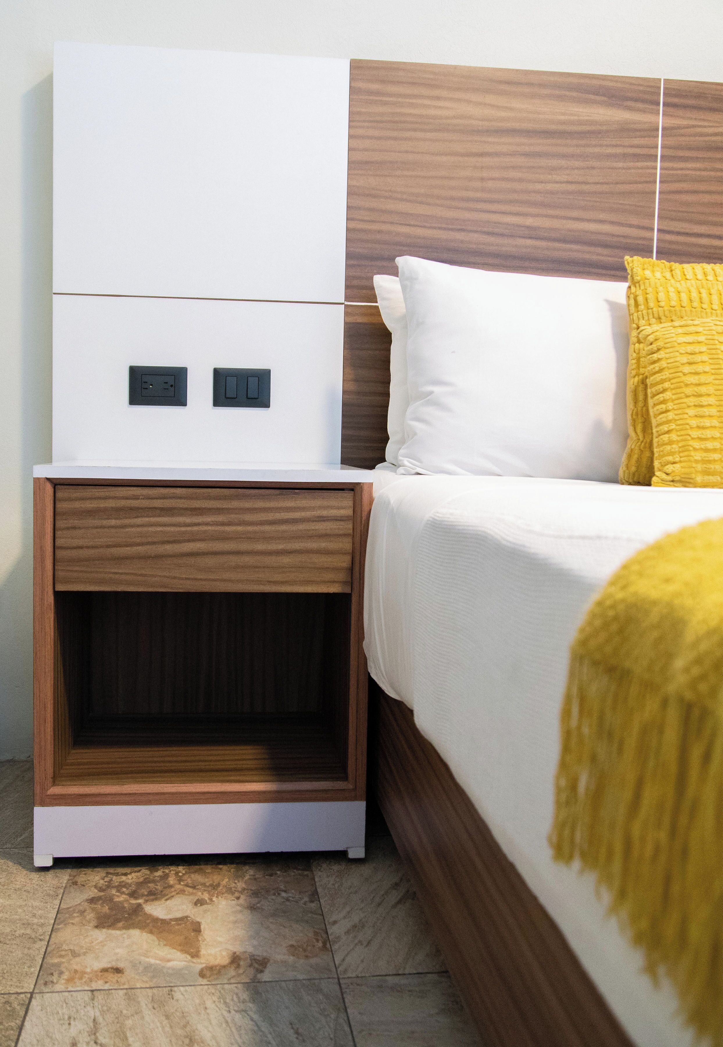 Een slaapkamer met een houten hoofdbord, witte beddengoed, een gele plaid en een nachtkastje met een lade.