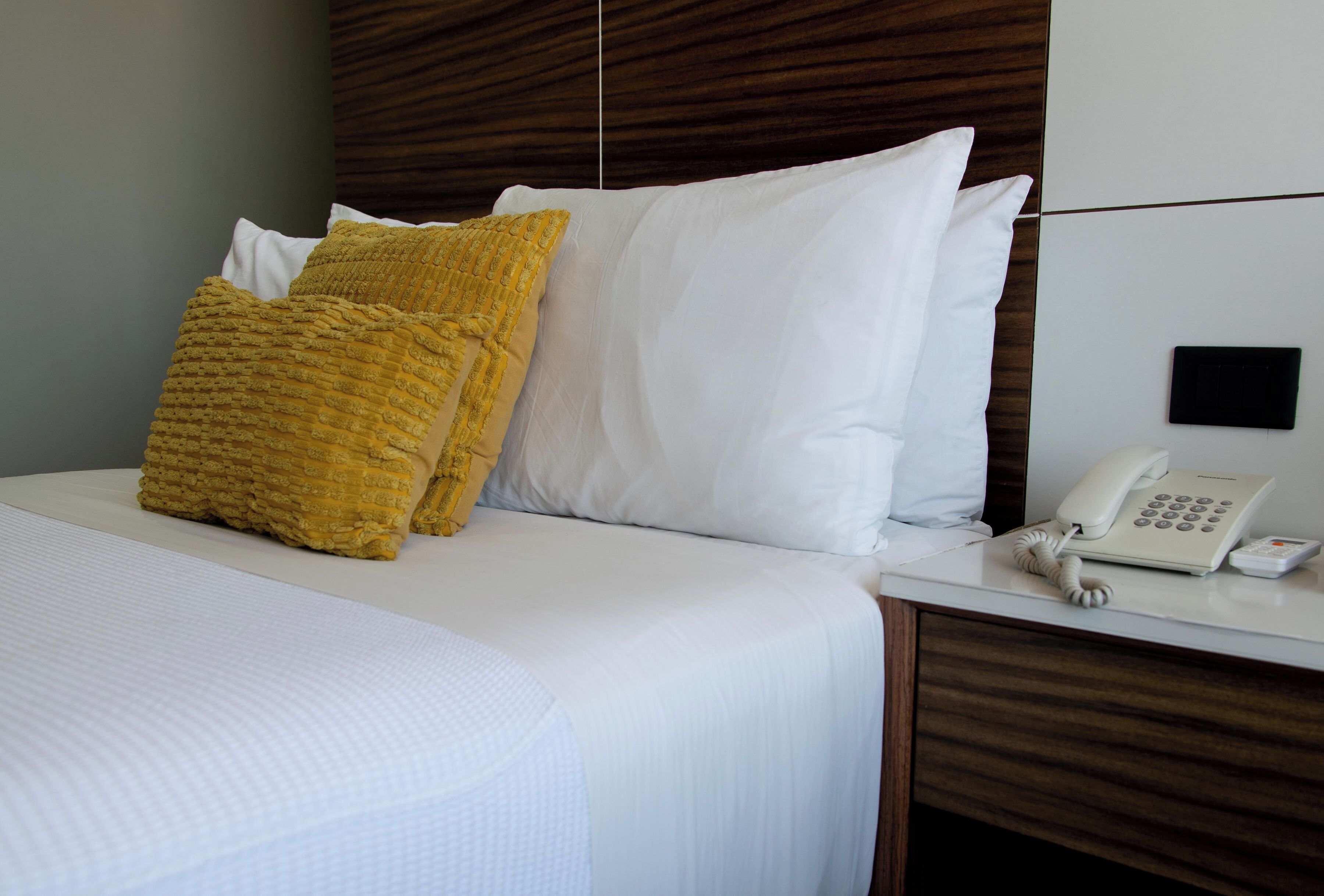 A neatly made bed with white and mustard yellow pillows, a white bedside table with a telephone, and a wooden headboard.