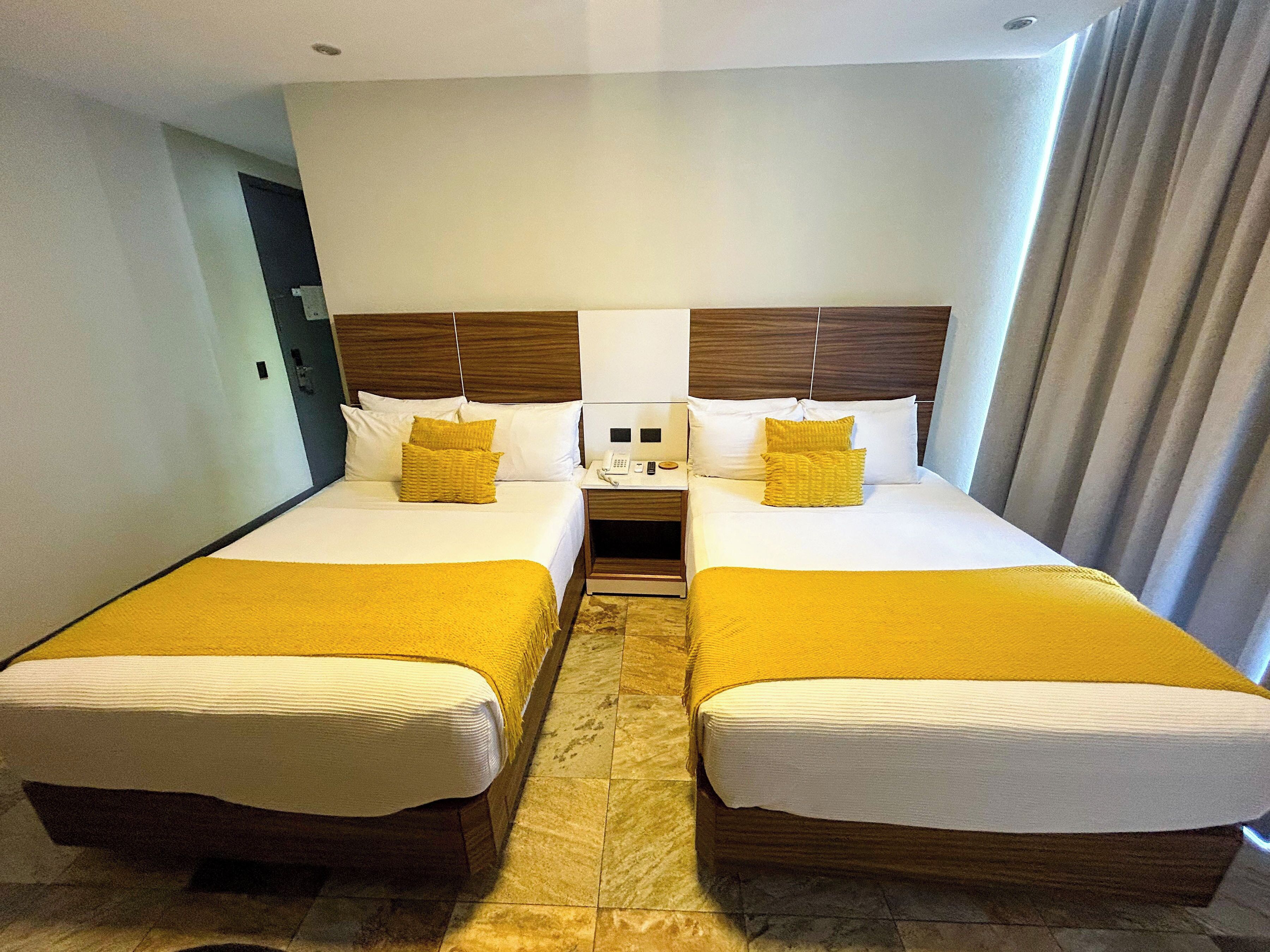 A hotel room with two beds, each with a wooden headboard, white bedding, and a yellow throw blanket. There is a bedside table with a phone and a lamp between the beds.
