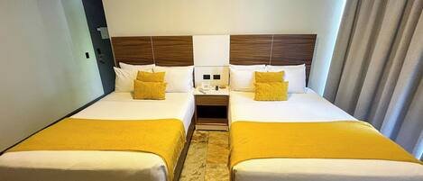 A hotel room with two beds, each with a wooden headboard, white bedding, and a yellow throw blanket. There is a bedside table with a phone and a lamp between the beds.
