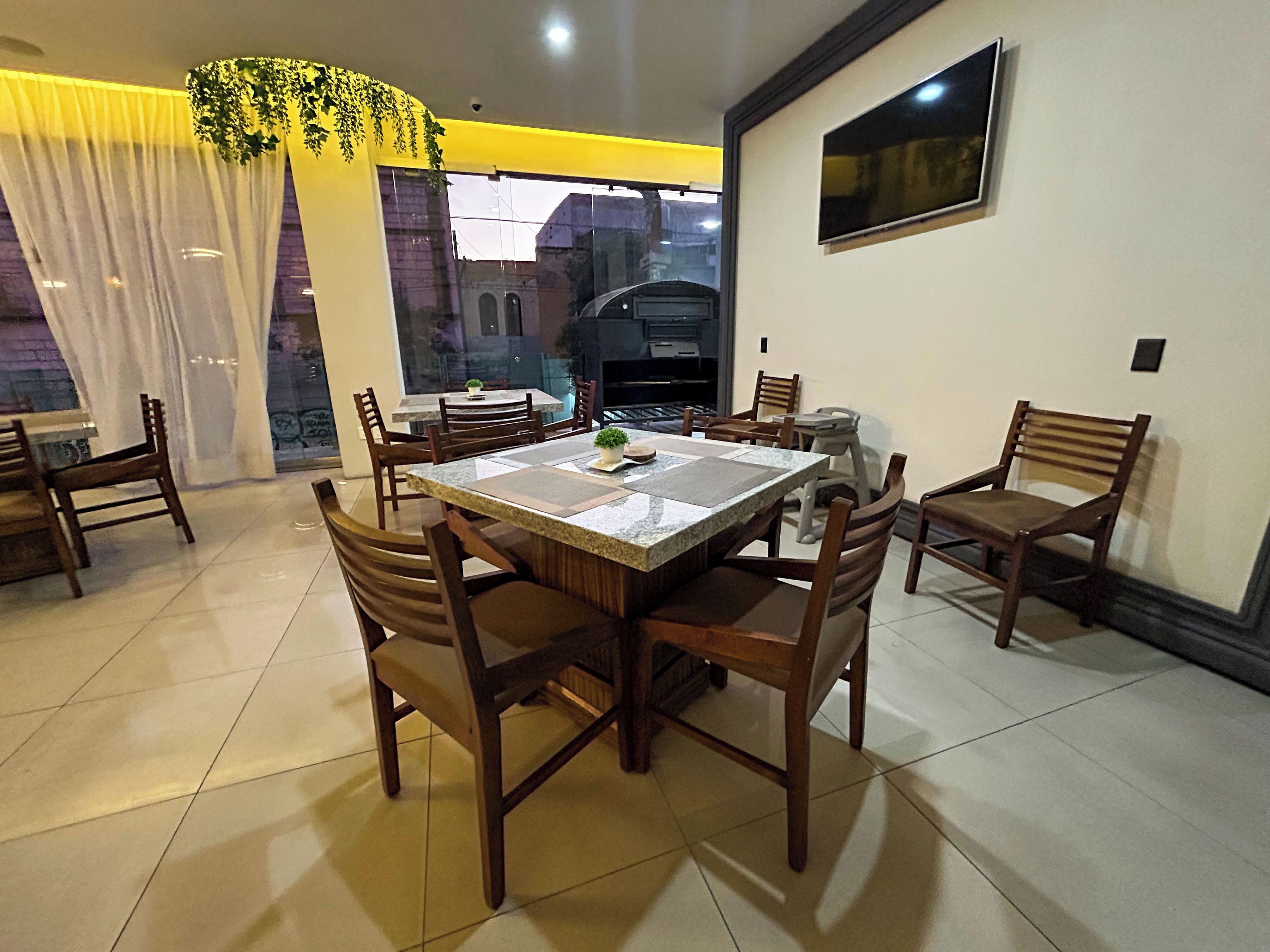 Un comedor con una mesa grande, varias sillas, un televisor montado en la pared y vistas de un edificio a través de una ventana.