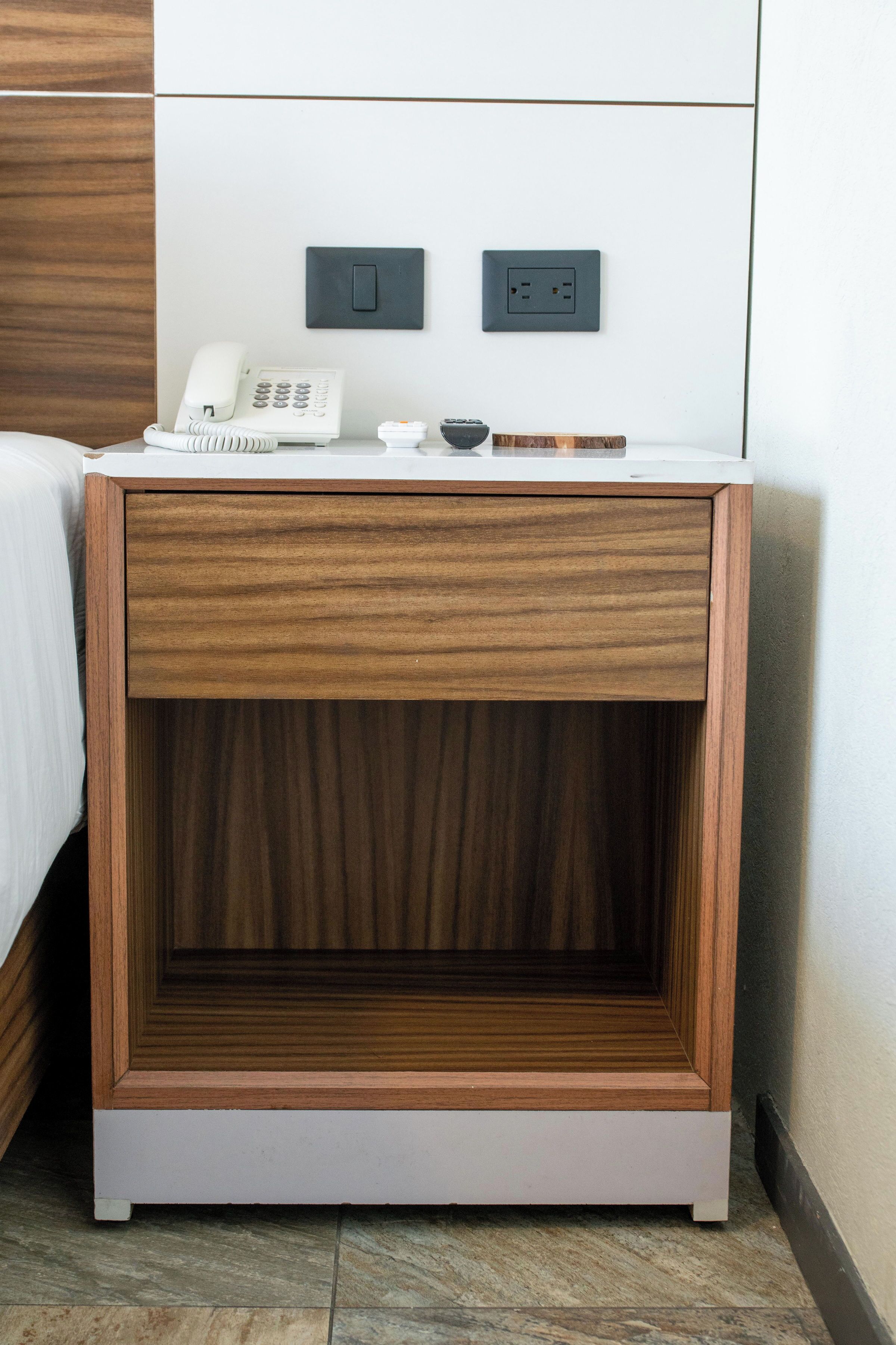 A wooden bedside table with a drawer, a white phone, and a small black object on top.
