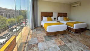 A hotel room with two beds, a large window, and a view of the street. - HVH by Yaxché (Guadalajara)
