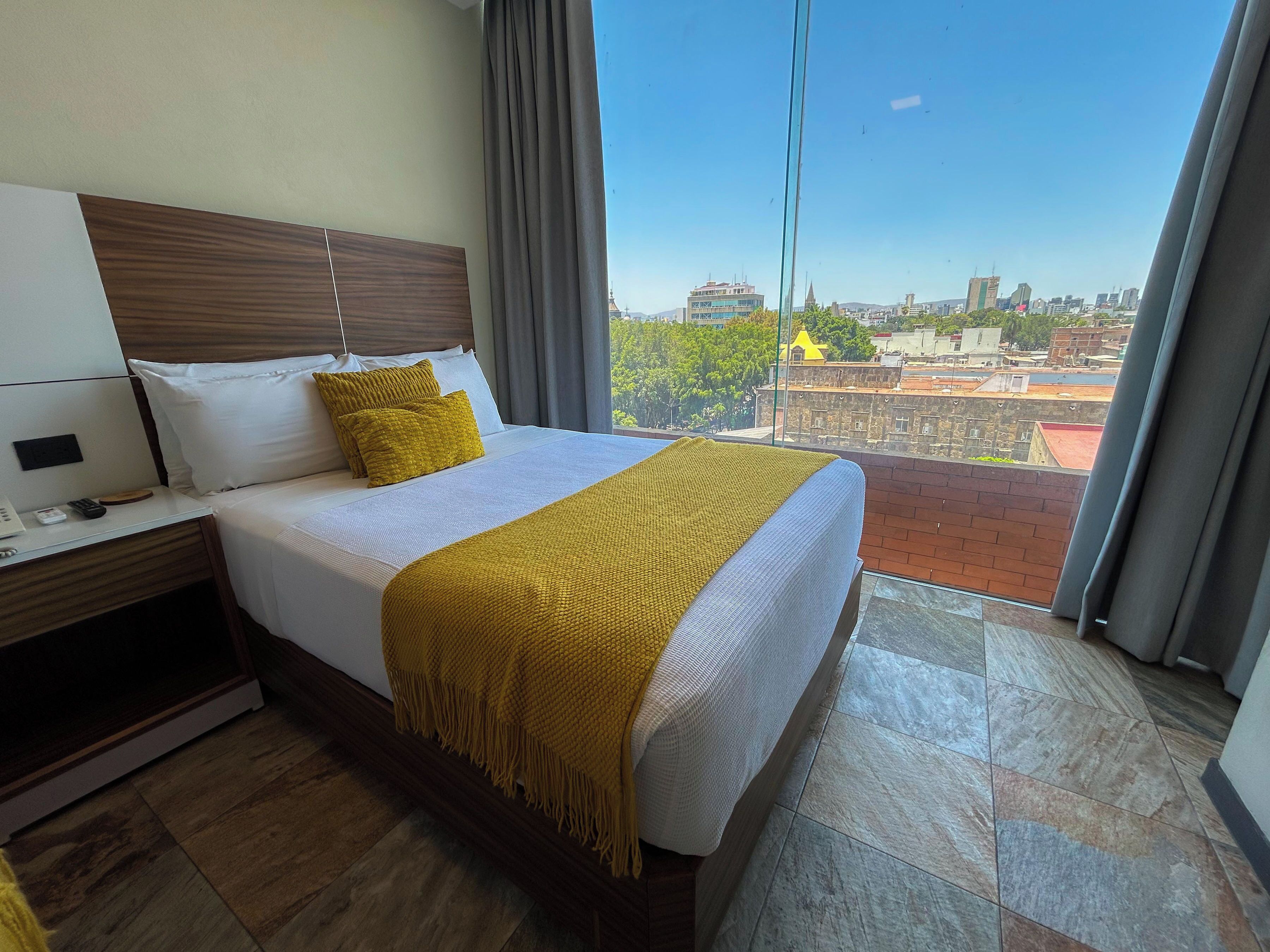 A hotel room with a bed, a nightstand, a window with a view of the city, and a balcony.