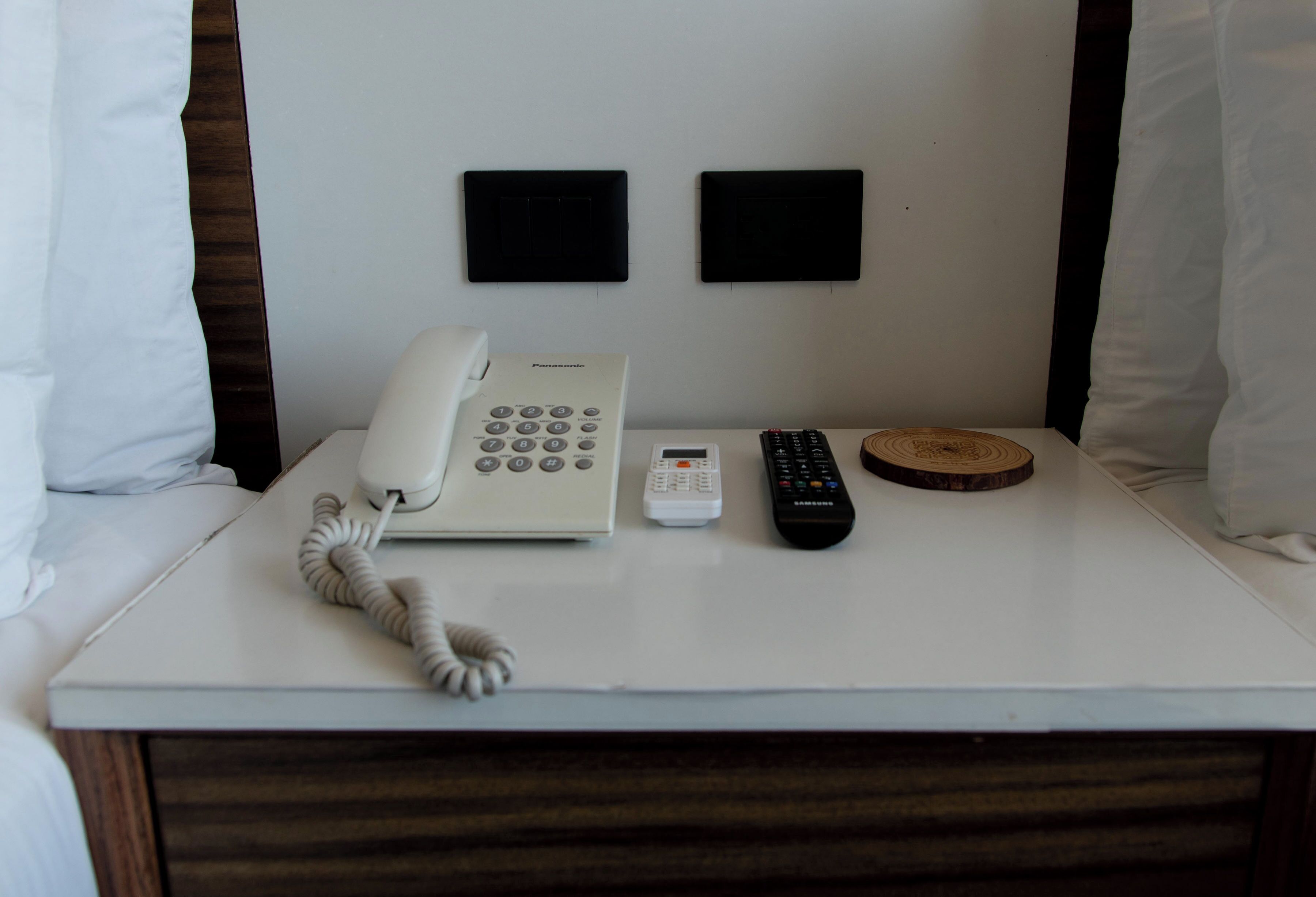 A hotel room with a white bedside table, a white telephone, a Samsung remote, a cordless phone, and two black wall-mounted air vents.