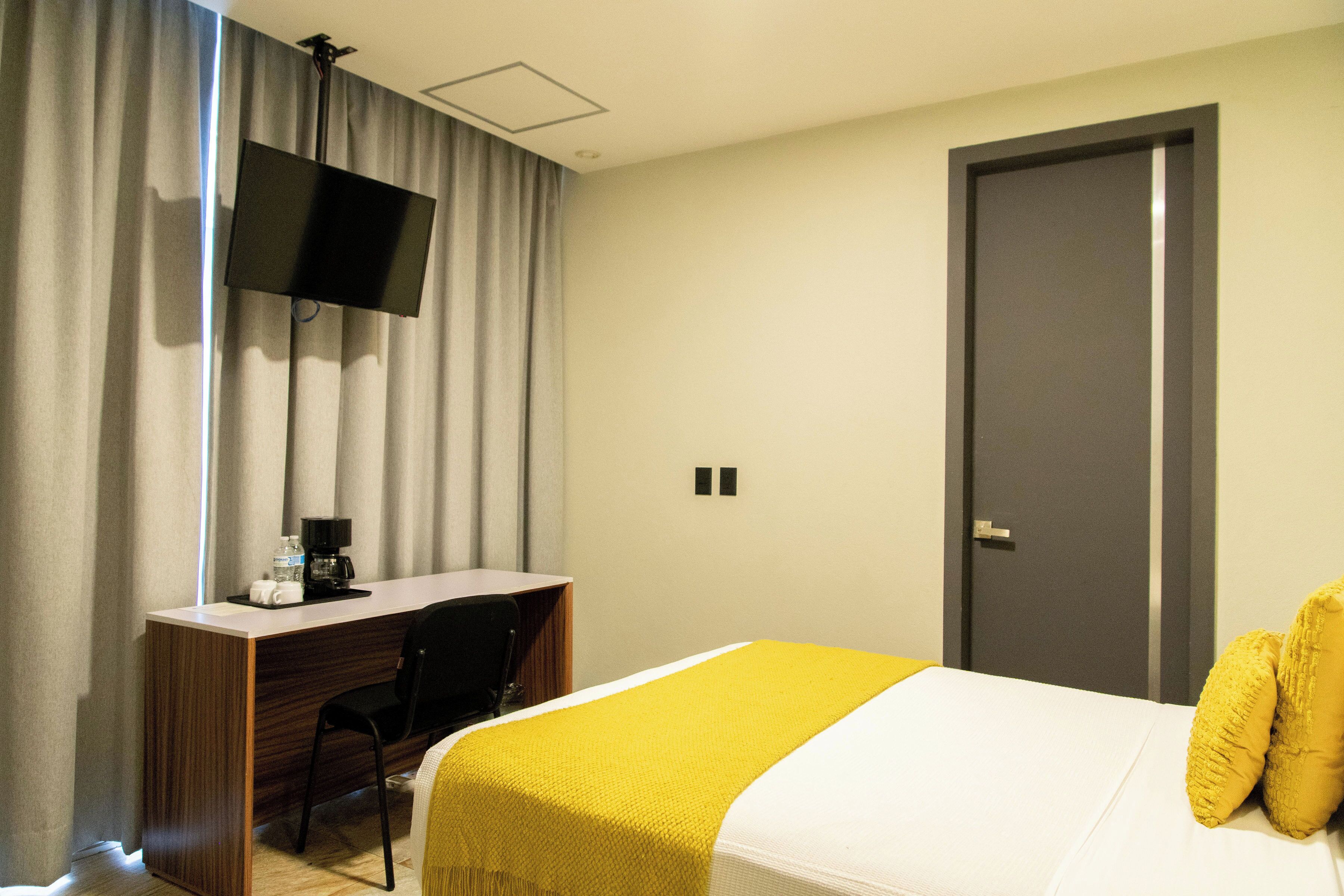 A hotel room with a bed, a desk with a chair, a television mounted on the wall, and a door.