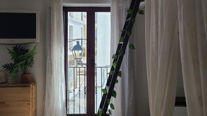A room with a tiled floor, a wooden dresser, a potted plant, a window with curtains, and a view of a street outside. - 113 Livingtarifa Amapola (Tarifa)