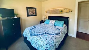 A bedroom with a bed, a dresser, a television, a framed picture, and a surfboard decoration on the wall.