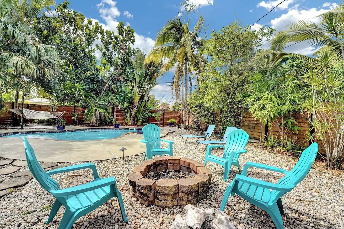 Un patio trasero con piscina, rodeado de plantas tropicales y una chimenea con sillas.